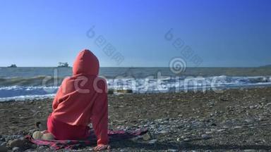 后景。 有人穿着红色运动衫和引擎盖在暴风雨中坐在海滩上看着大海